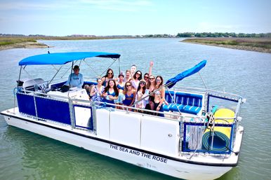 Happy group of friends on a blue-and-white pontoon boat with canopy cruising a calm coastal marsh channel, posing with drinks on a sunny day.