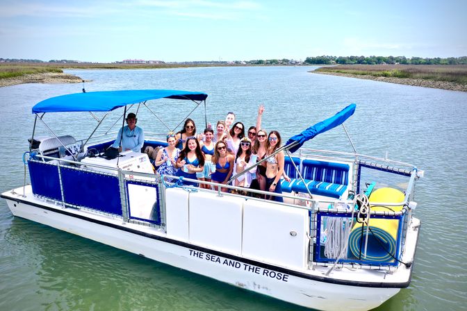 Happy group of friends on a blue-and-white pontoon boat with canopy cruising a calm coastal marsh channel, posing with drinks on a sunny day.