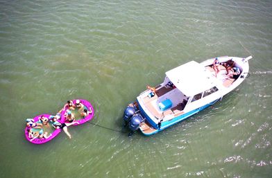 Aerial view of a blue-and-white motorboat in green water towing two connected pink party floats with people lounging and sunbathing.
