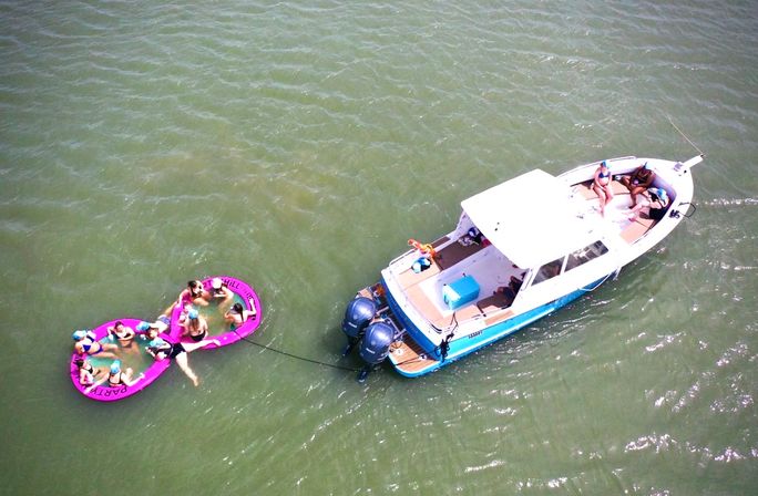 Aerial view of a blue-and-white motorboat in green water towing two connected pink party floats with people lounging and sunbathing.