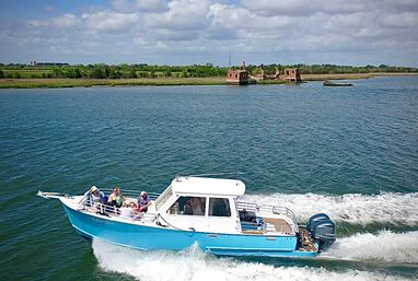 Blue-and-white motorboat carrying passengers zips across a calm bay, leaving a white wake against deep blue water with a grassy coastal marsh and small abandoned brick structures on the distant shore under a partly cloudy sky.