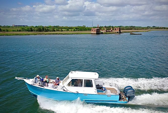 Blue-and-white motorboat carrying passengers zips across a calm bay, leaving a white wake against deep blue water with a grassy coastal marsh and small abandoned brick structures on the distant shore under a partly cloudy sky.