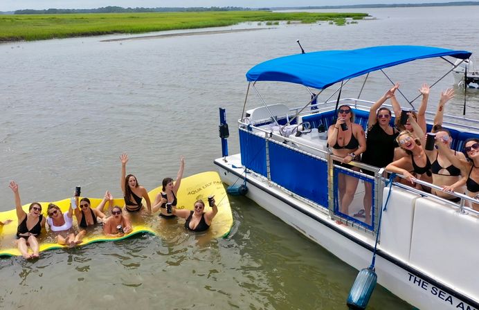 Group of women in swimsuits cheering on a bright yellow floating mat beside a blue-canopy pontoon boat in a calm coastal salt‑marsh estuary, summer boat party vibe.