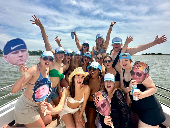Cheerful group of friends in swimsuits and caps on a sunny lake boat, laughing, raising hands and holding oversized face cutouts and drinks.