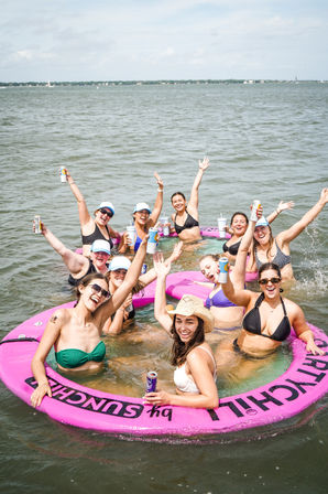 Smiling group of women in swimsuits cheering and holding drinks while relaxing in a large pink inflatable float on a sunny bay.