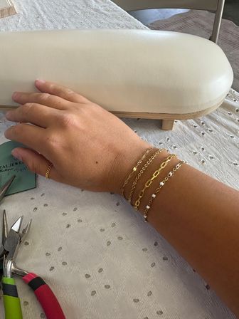 Close-up: hand wearing stacked delicate gold chain bracelets and a thin gold ring on a jewelry bench beside pliers and a white padded display pillow on a textured cloth