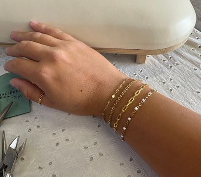 Close-up: hand wearing stacked delicate gold chain bracelets and a thin gold ring on a jewelry bench beside pliers and a white padded display pillow on a textured cloth
