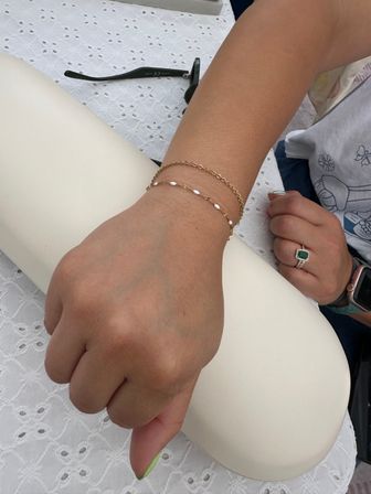Close-up of a person’s hand on a padded armrest showcasing two delicate gold chain bracelets with small white beads, green-tipped manicure, emerald halo ring and smartwatch nearby on a white eyelet tablecloth with sunglasses in the background.