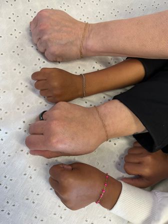 Four diverse wrists resting on white eyelet fabric, each wearing dainty bracelets — thin gold chains, a silver chain and a pink beaded bracelet — showcasing mixed skin tones and minimalist jewelry.