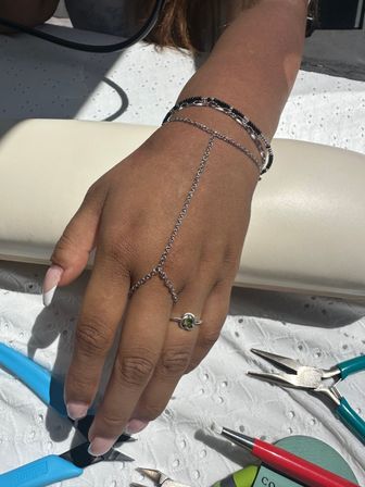 Sunlit hand wearing a delicate silver hand-chain bracelet and green-stone ring resting on a white mat in an outdoor jewelry workspace with pliers and tools