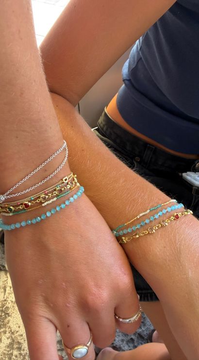 Close-up of two sunlit wrists wearing stacked turquoise bead, gold and silver chain bracelets with small gemstone accents and rings, casual navy top and black denim visible.