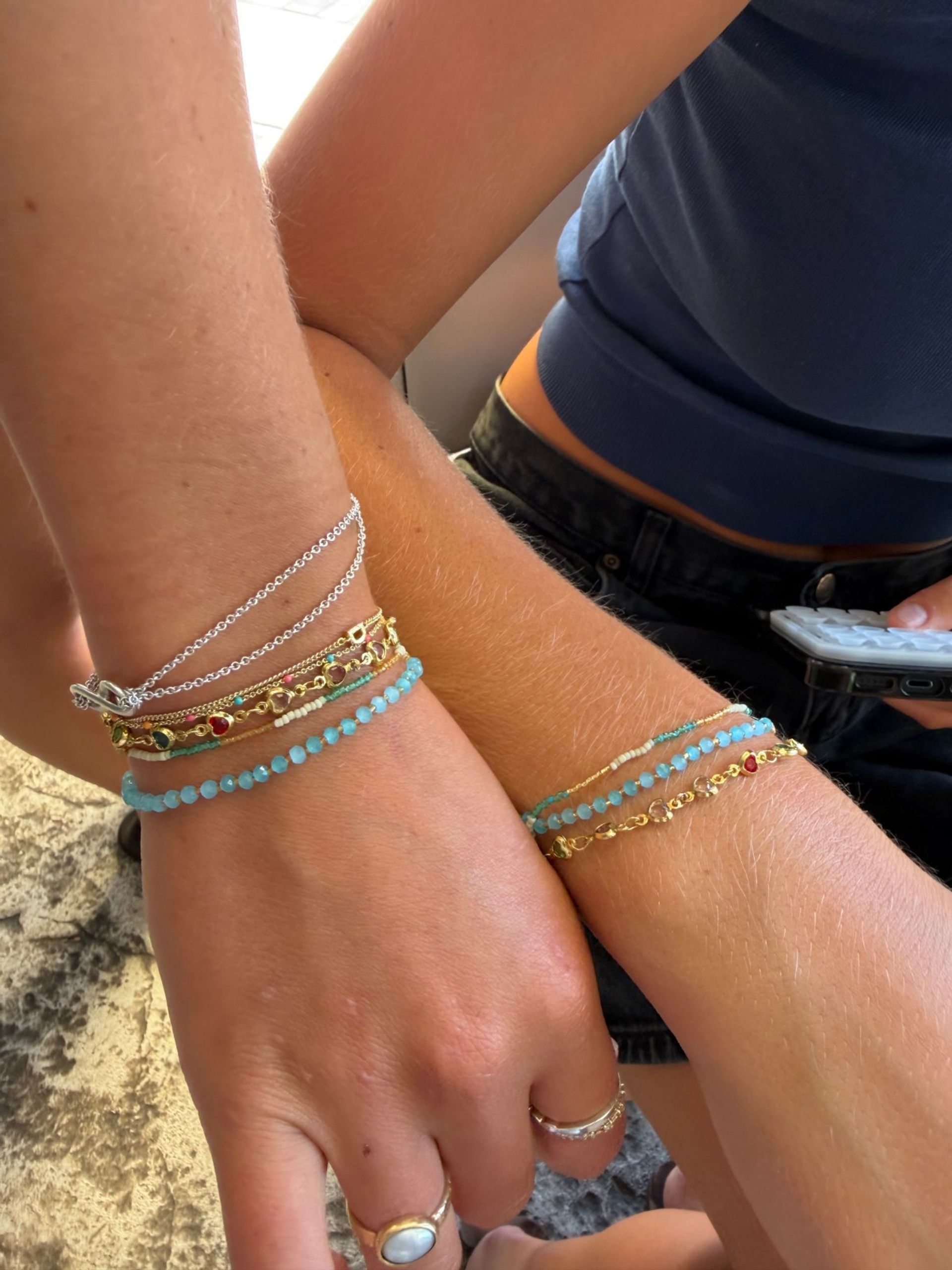 Close-up of two sunlit wrists wearing stacked turquoise bead, gold and silver chain bracelets with small gemstone accents and rings, casual navy top and black denim visible.
