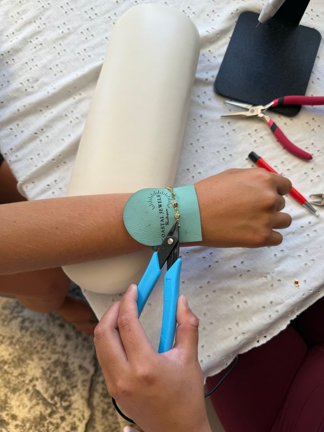 Close-up of DIY jewelry-making: hands use blue pliers to clasp a delicate gold beaded bracelet on a forearm resting on a white wrist cushion and teal leather pad, with red-handled tools on a cloth-covered table.