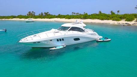 White luxury motor yacht anchored in crystal turquoise water off a sandy tropical island beach with palm trees, paddleboard and jet ski tied alongside and smaller boats in the background.