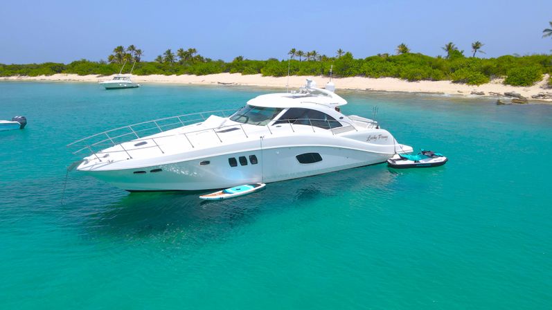White luxury motor yacht anchored in crystal turquoise water off a sandy tropical island beach with palm trees, paddleboard and jet ski tied alongside and smaller boats in the background.