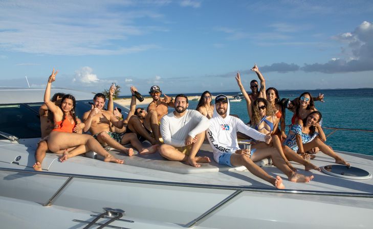 Group of friends lounging on the foredeck of a white yacht, cheering with drinks and posing in swimsuits on a sunny day over turquoise tropical waters with a sandy island in the background.