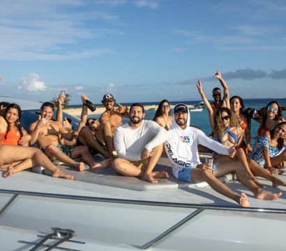 Group of friends lounging on the foredeck of a white yacht, cheering with drinks and posing in swimsuits on a sunny day over turquoise tropical waters with a sandy island in the background.
