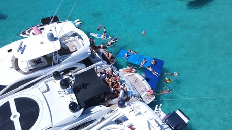 Aerial view of luxury yachts clustered in crystal-clear turquoise water, sun-soaked boat party with people lounging on decks and blue floating mats, swimmers and paddleboarders nearby.