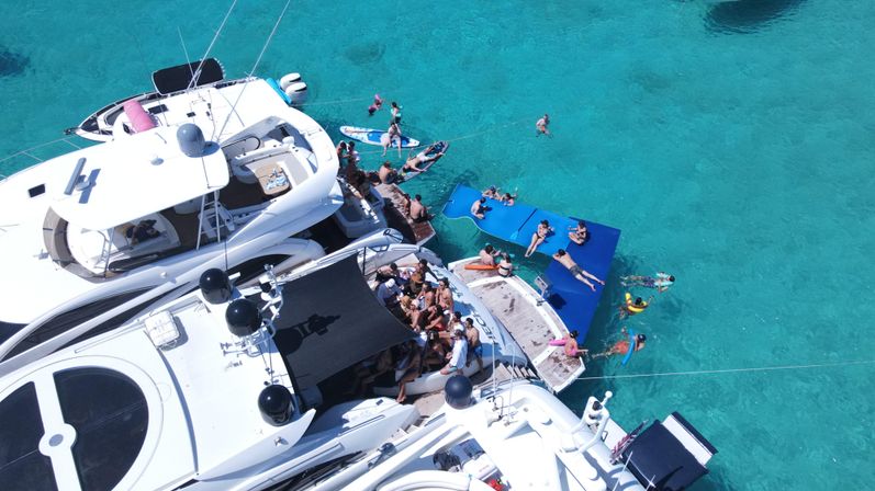 Aerial view of luxury yachts clustered in crystal-clear turquoise water, sun-soaked boat party with people lounging on decks and blue floating mats, swimmers and paddleboarders nearby.