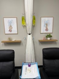 Infusion clinic treatment room with two black reclining chairs flanking a curtained station, two bright yellow IV bags hanging, framed syringe artwork and shelves, and a prep tray with medical supplies.