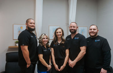 Five smiling healthcare professionals in black scrubs posing for a clinic team portrait in a bright treatment room with neutral decor and framed art