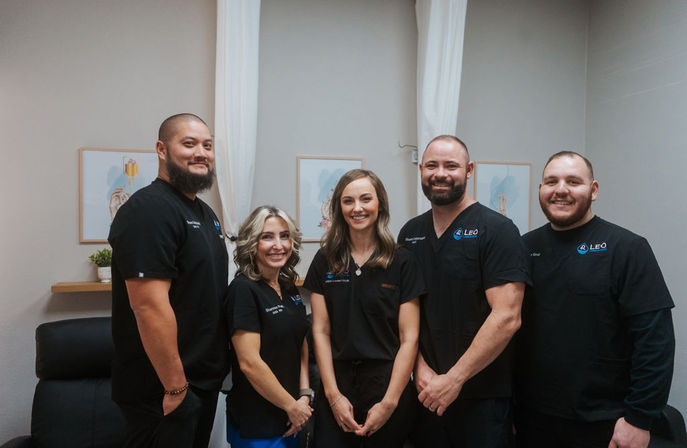 Five smiling healthcare professionals in black scrubs posing for a clinic team portrait in a bright treatment room with neutral decor and framed art