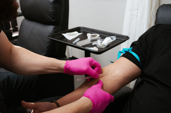 Phlebotomist in bright pink gloves performing a routine blood draw from a patient’s arm with a blue tourniquet in a medical clinic setting