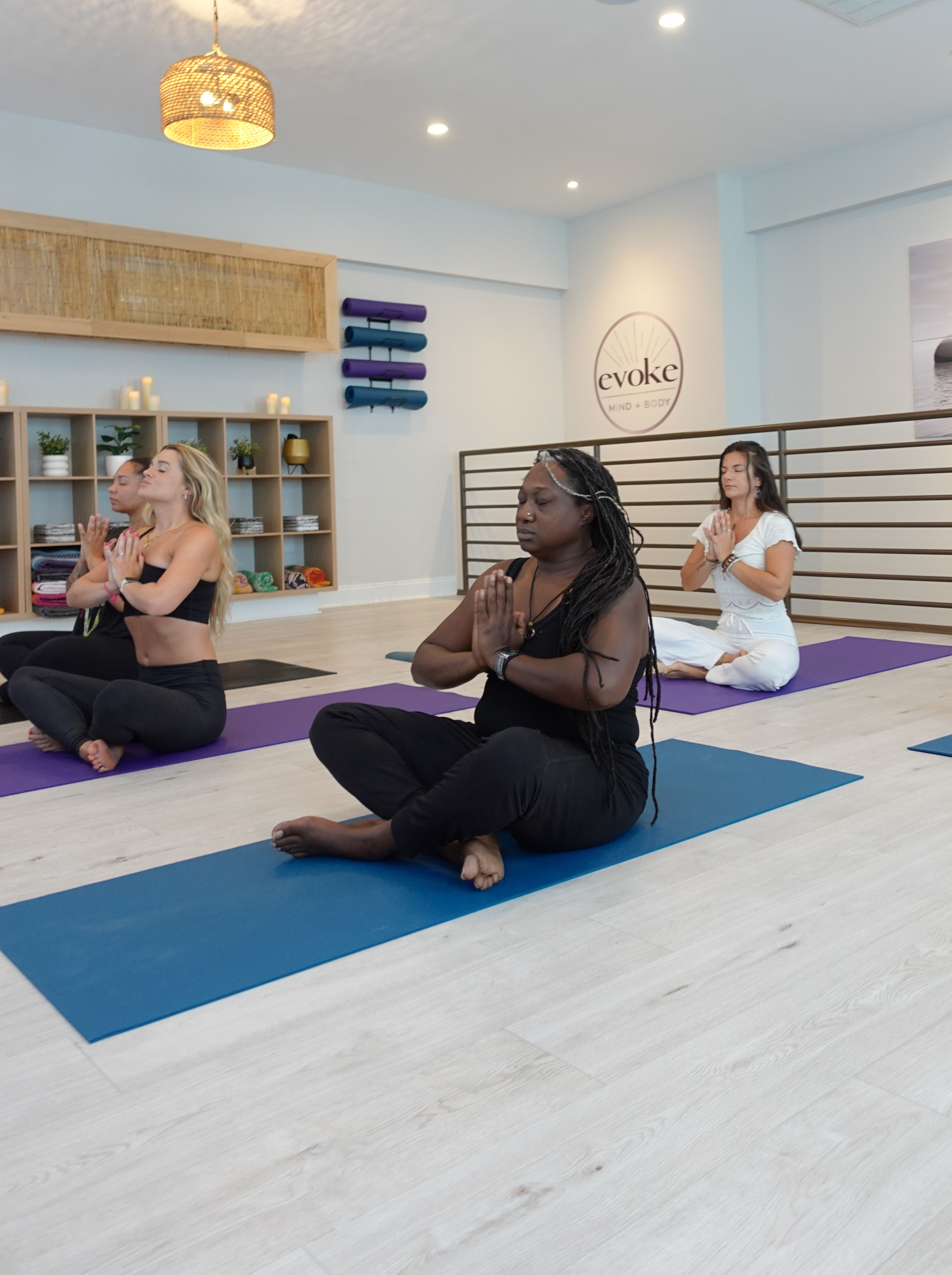 Women in a bright yoga studio seated cross‑legged on colorful mats with hands in prayer pose, practicing calm meditation amid stacked mats, shelves and soft lighting.
