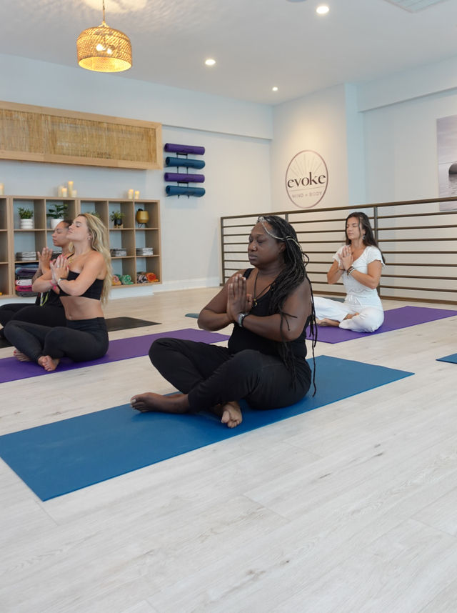 Women in a bright yoga studio seated cross‑legged on colorful mats with hands in prayer pose, practicing calm meditation amid stacked mats, shelves and soft lighting.