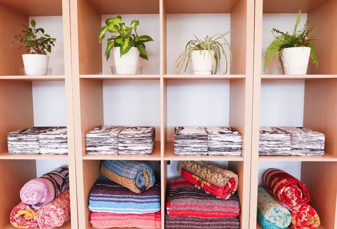 Wooden cubby shelving neatly arranged with potted indoor plants on top, stacks of folded patterned linens in the middle, and colorful rolled blankets and throws on the bottom — bright, organized home decor display.