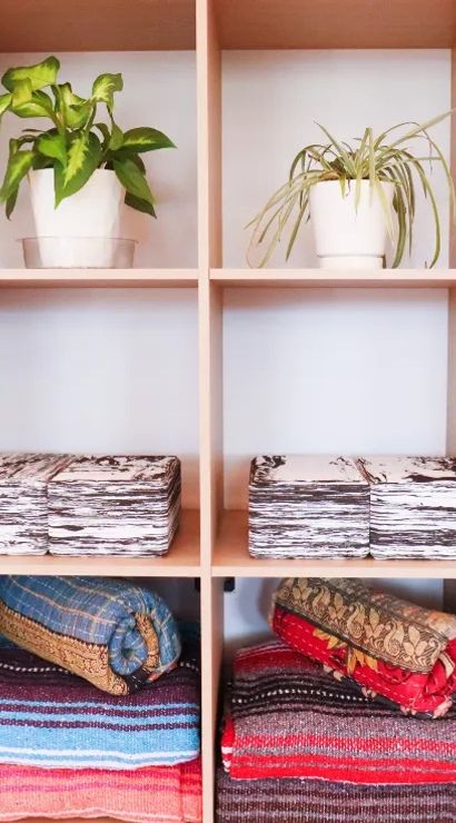 Light wood cubby shelves showing a bright home-decor display: white pots with green houseplants on the top row, stacks of black-and-white folded textiles in the middle, and colorful rolled blankets and woven throws on the bottom.