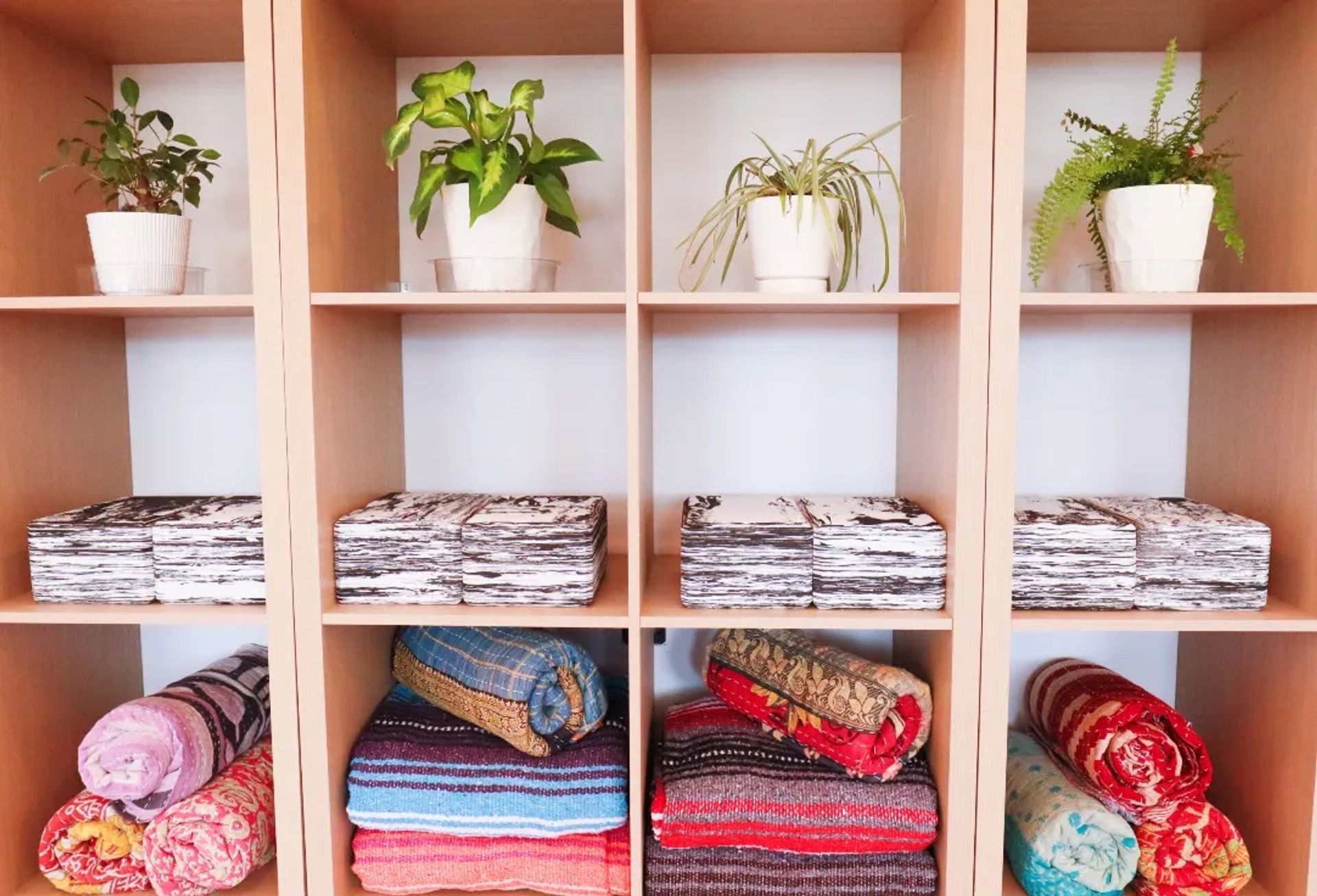 Light wood cubby shelves showing a bright home-decor display: white pots with green houseplants on the top row, stacks of black-and-white folded textiles in the middle, and colorful rolled blankets and woven throws on the bottom.
