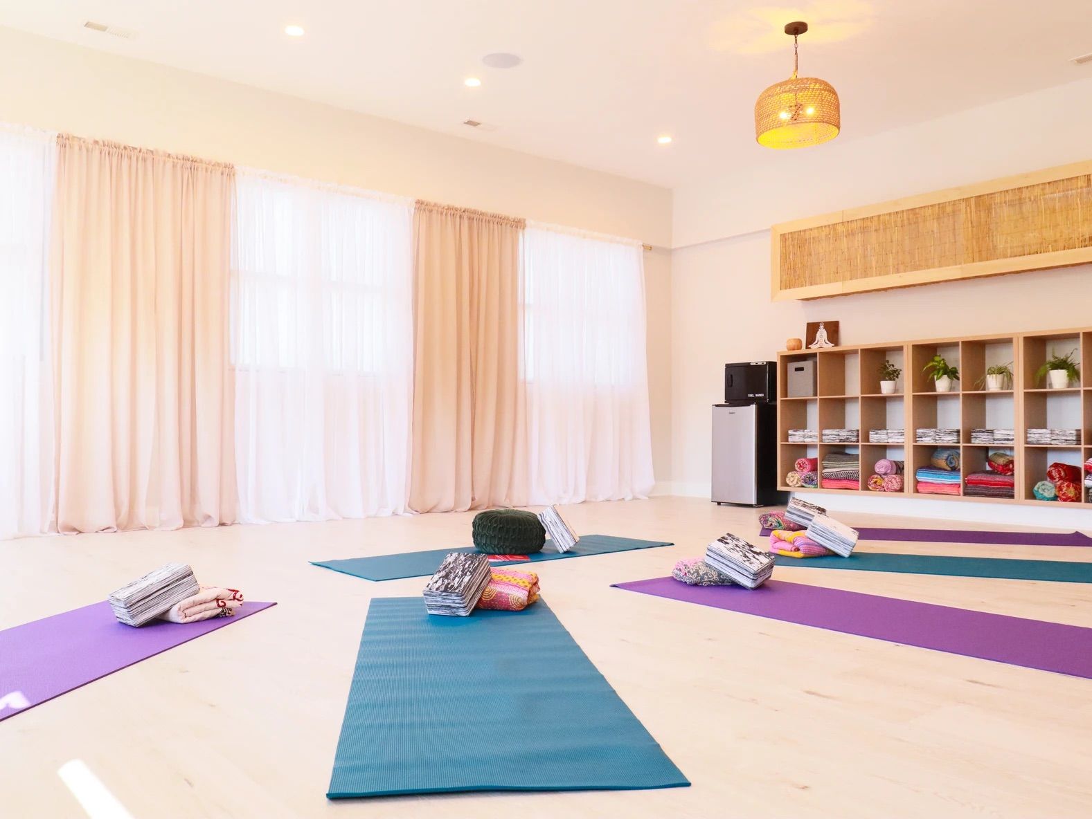 Bright airy yoga studio interior with teal and purple mats, bolsters and folded blankets on cubby shelving, potted plants and soft floor-to-ceiling curtains