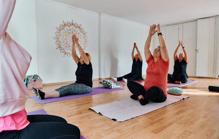 Senior women in a community yoga class in a bright indoor studio, seated on mats on a wooden floor reaching arms overhead beneath a decorative mandala wall art