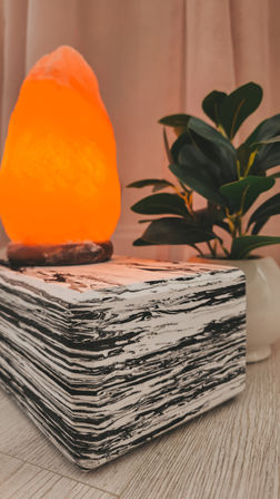 Glowing orange salt lamp on a black-and-white marbled cube table beside a leafy potted plant — cozy indoor home decor vignette.