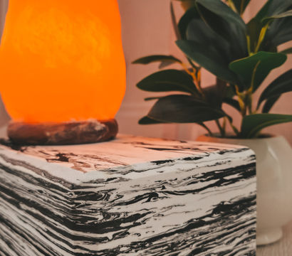 Glowing orange salt lamp on a black-and-white marbled cube table beside a leafy potted plant — cozy indoor home decor vignette.
