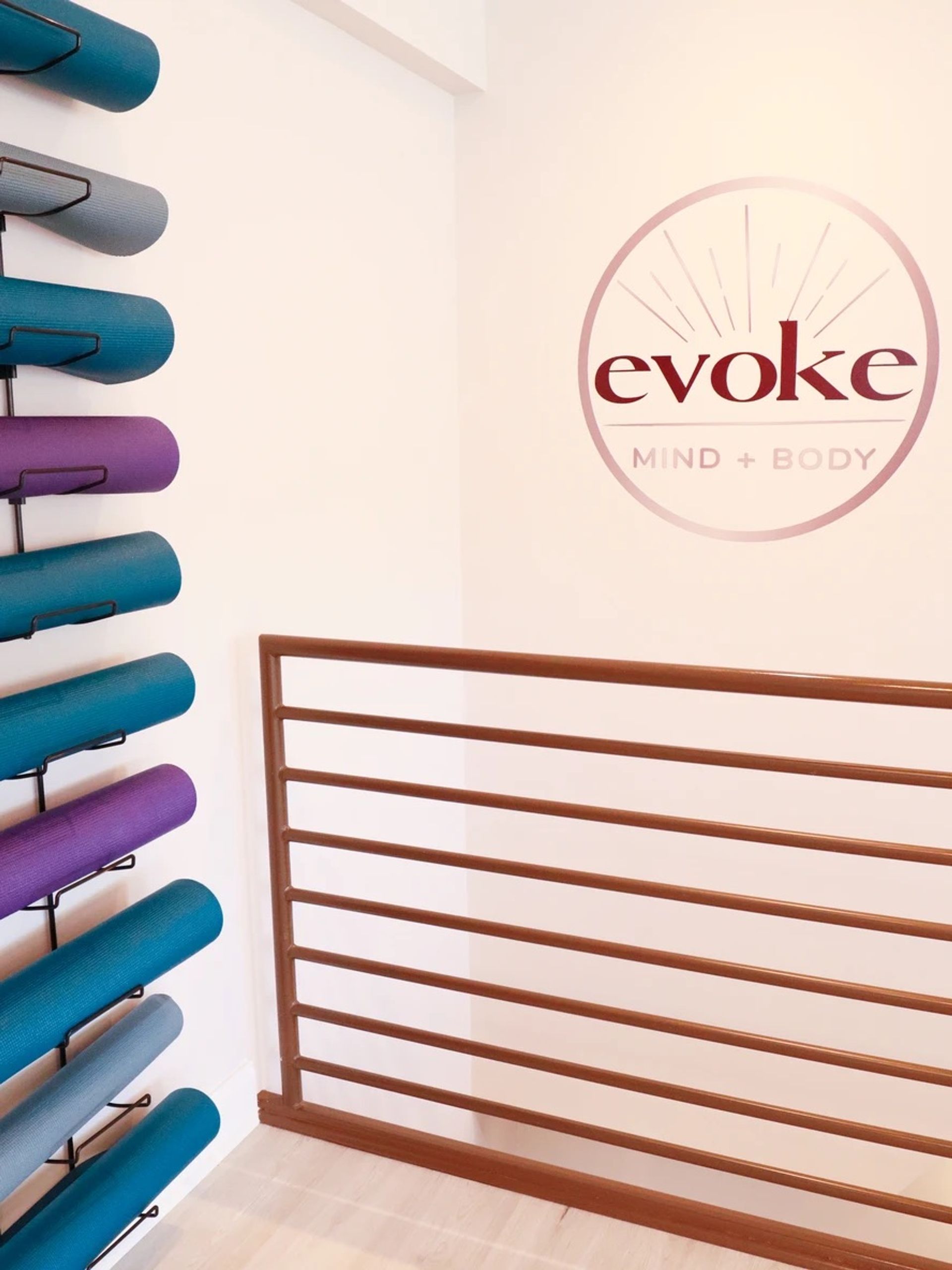 Bright wellness studio corner with teal, purple and gray yoga mats on a vertical wall rack, wooden horizontal railing and a circular wall logo on a pale wall, indoor yoga/fitness studio interior.