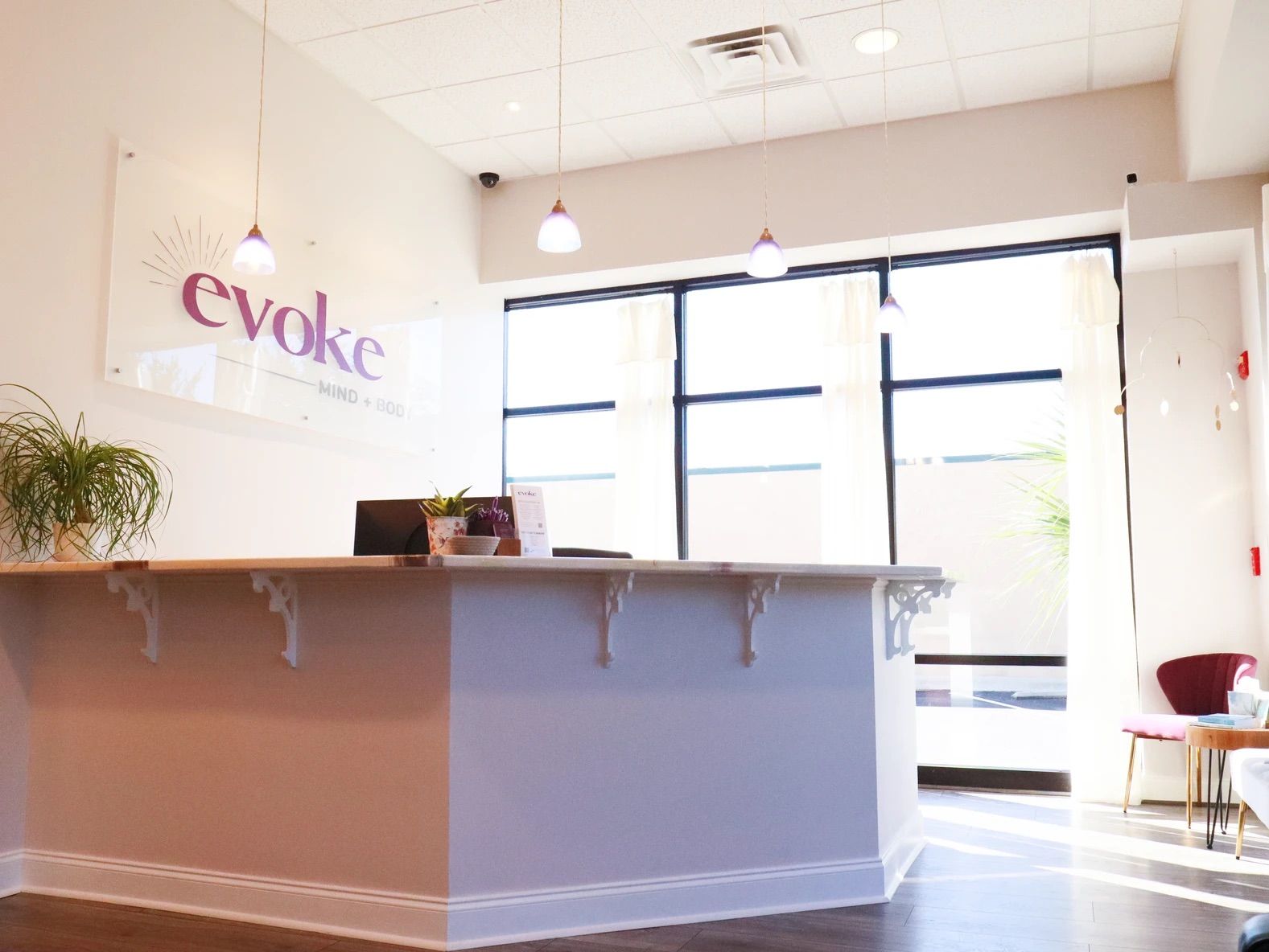 Sunlit modern wellness studio reception with a white front desk, hanging pendant lights, large windows, potted plants and cozy seating