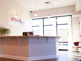 Sunlit modern wellness studio reception with a white front desk, hanging pendant lights, large windows, potted plants and cozy seating
