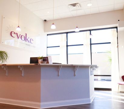 Sunlit modern wellness studio reception with a white front desk, hanging pendant lights, large windows, potted plants and cozy seating
