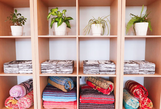 Light wood cubby shelf styled for home decor: potted houseplants on top, marble-patterned storage boxes in the middle, and colorful folded blankets and rolled bolsters on the bottom.