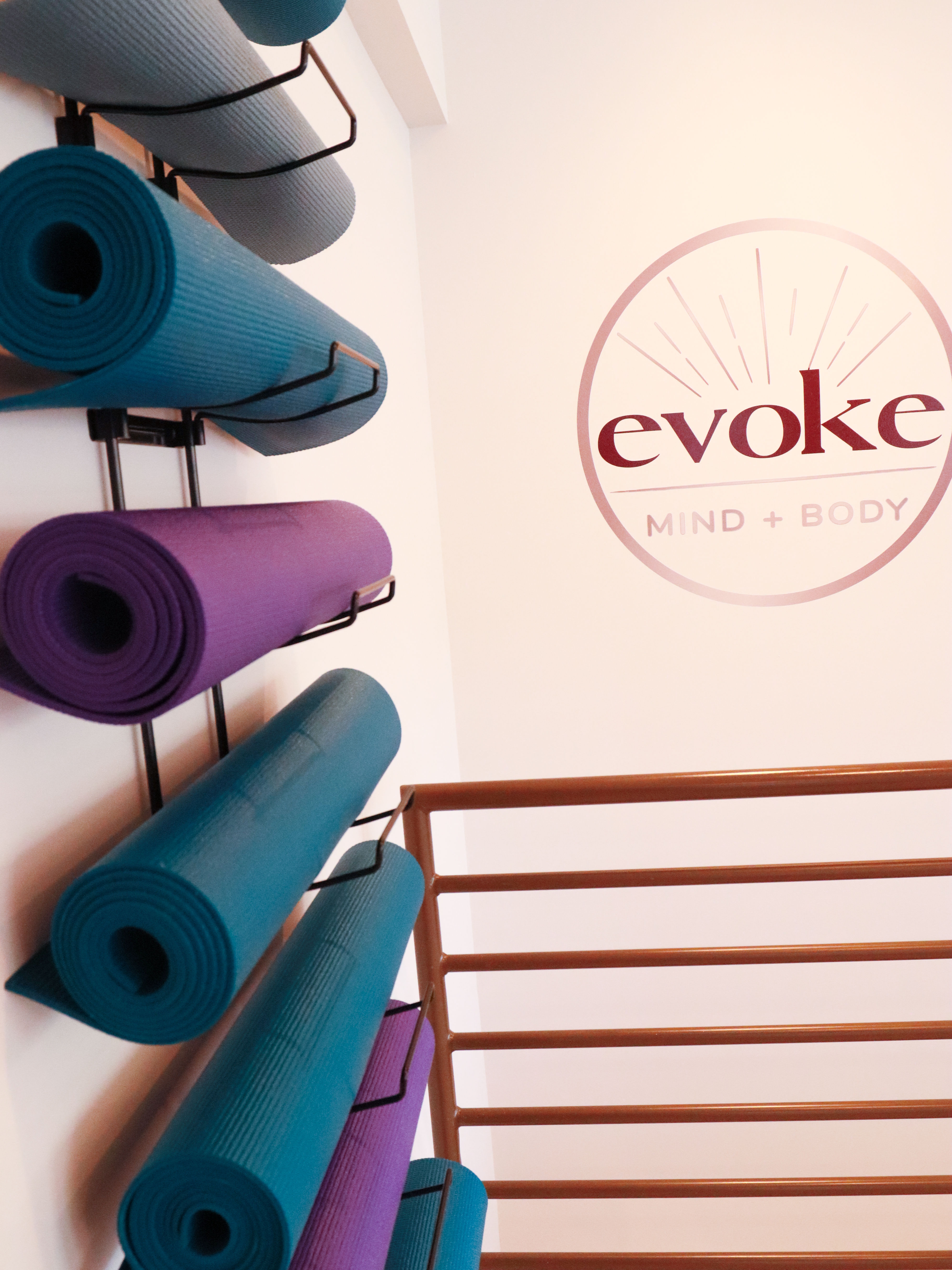 Rolled teal and purple yoga mats neatly stored on a wall rack next to a wellness studio logo and wooden barre — bright, minimalist yoga/fitness studio interior.