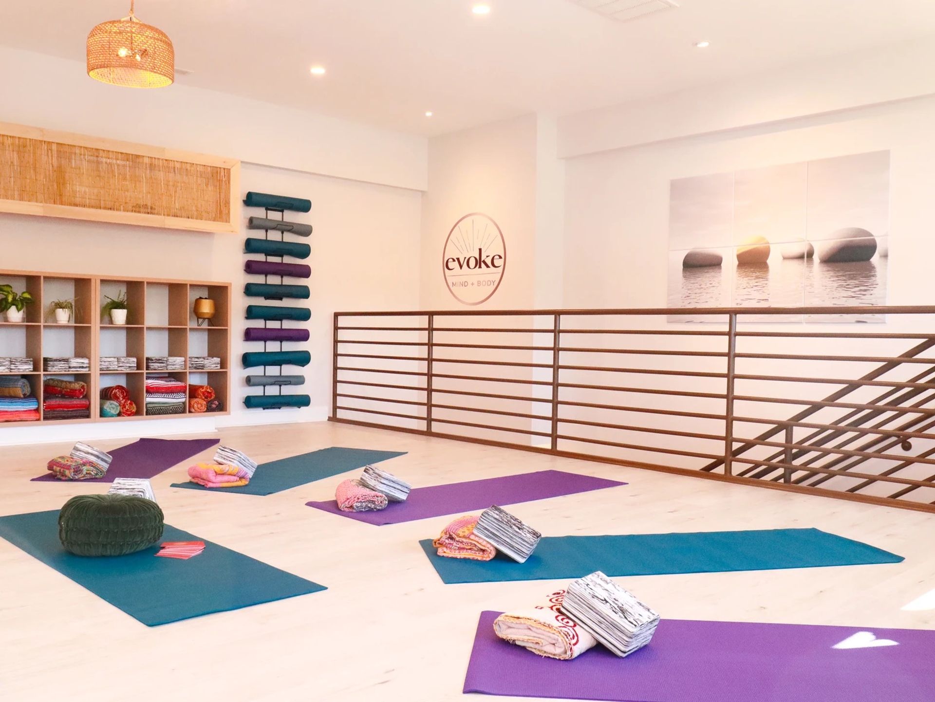 Sunlit, minimalist yoga studio interior with colorful mats and bolsters on a light wood floor, cubby shelves of blankets and plants, stacked wall mats, metal railing and calming stone wall art.