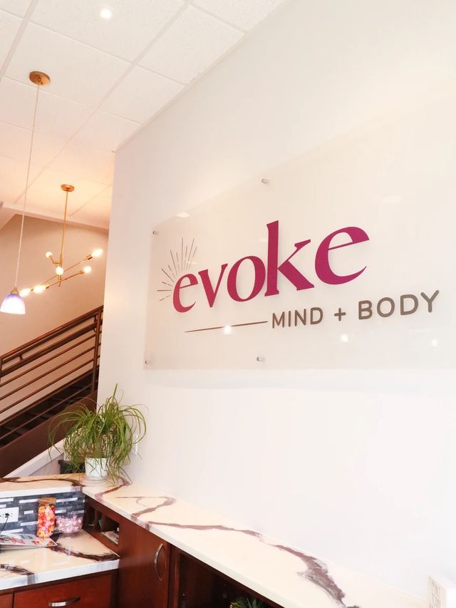 Bright modern spa reception — welcoming wellness studio lobby with oversized wall sign, marble-pattern countertop, potted plant, brass pendant lights, and a staircase.