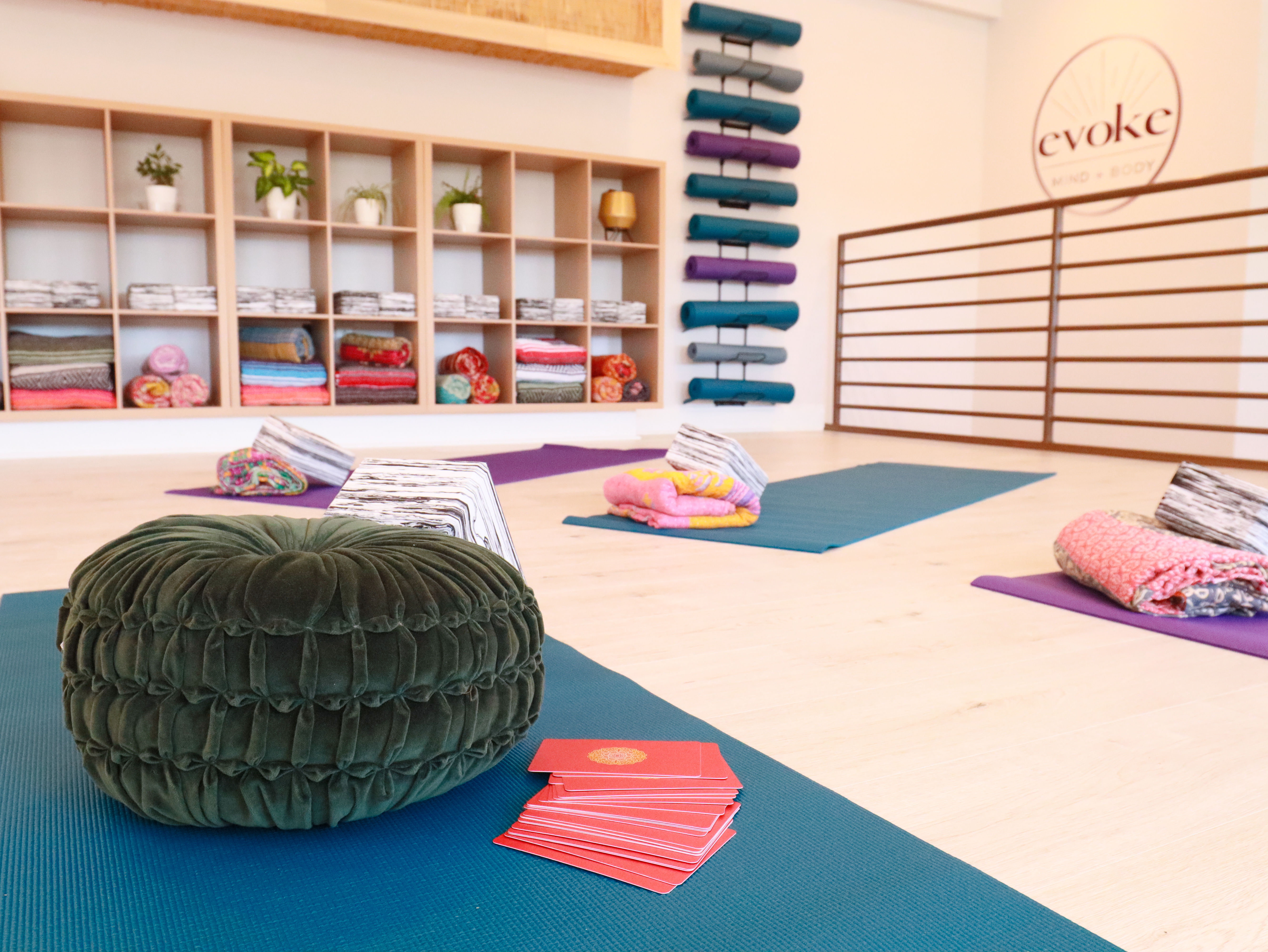 Bright modern yoga studio interior with teal mats, round meditation cushions, colorful folded blankets in cubby shelves and rolled yoga mats on a wall rack