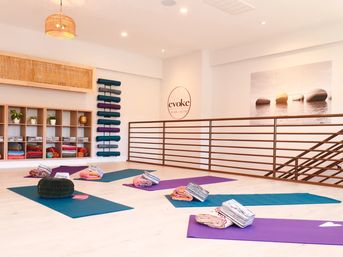 Sunlit modern yoga studio interior with colorful mats and rolled blankets neatly arranged on a pale wood floor, stacked bolsters and shelving with plants and props, minimalist railing and calming wall art.