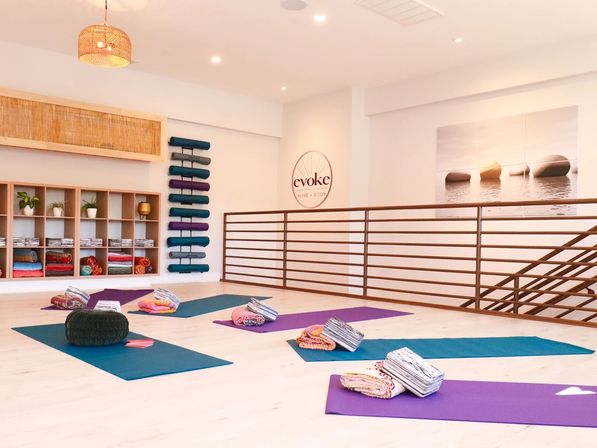 Sunlit modern yoga studio interior with colorful mats and rolled blankets neatly arranged on a pale wood floor, stacked bolsters and shelving with plants and props, minimalist railing and calming wall art.