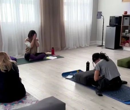 Sunlit indoor yoga studio with a small group seated on mats for a quiet restorative yoga and meditation session near sheer curtains, yoga props and water bottles.