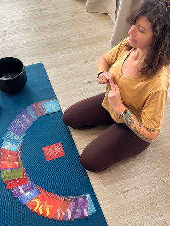 Person kneeling on a blue yoga mat indoors arranging a fan of colorful oracle/affirmation cards beside a black singing bowl, wearing a mustard top and showing a tattooed forearm — calm home meditation setup.