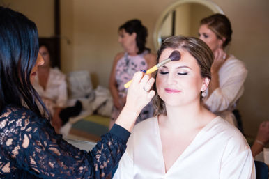 Bride in a satin robe smiling with eyes closed as a makeup artist applies powder with a large brush, bridesmaids and a mirror visible in the bridal suite during wedding morning preparations.